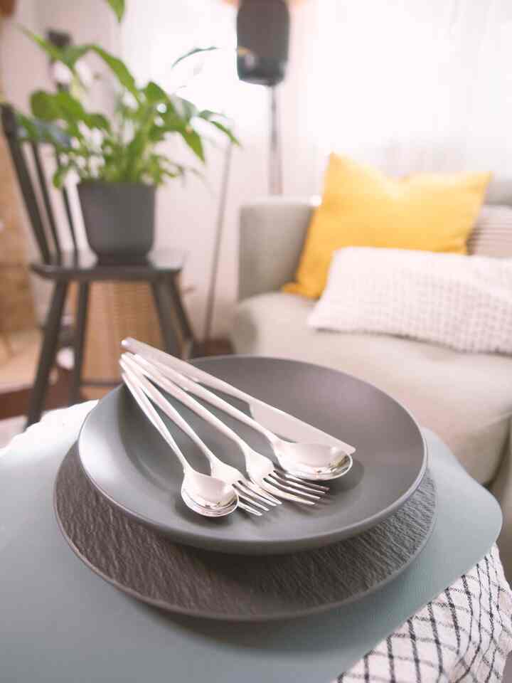 A bright white background living room with gray sofa and yellow cushion featuring matte black plates and silver cutlery arranged centrally, embodying a simple modern look