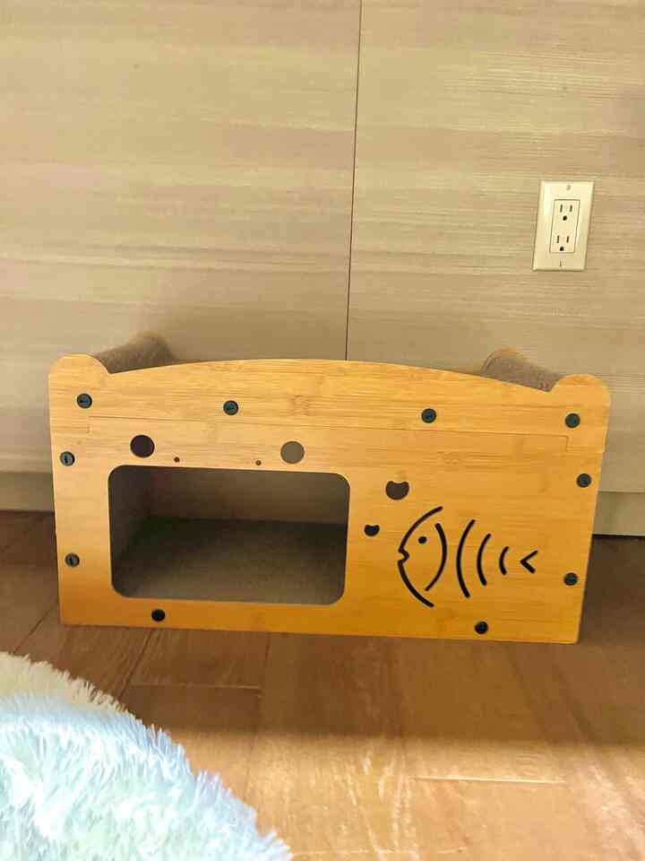 Natural wood-toned cozy corner featuring a scratch board cat house against a beige wall background