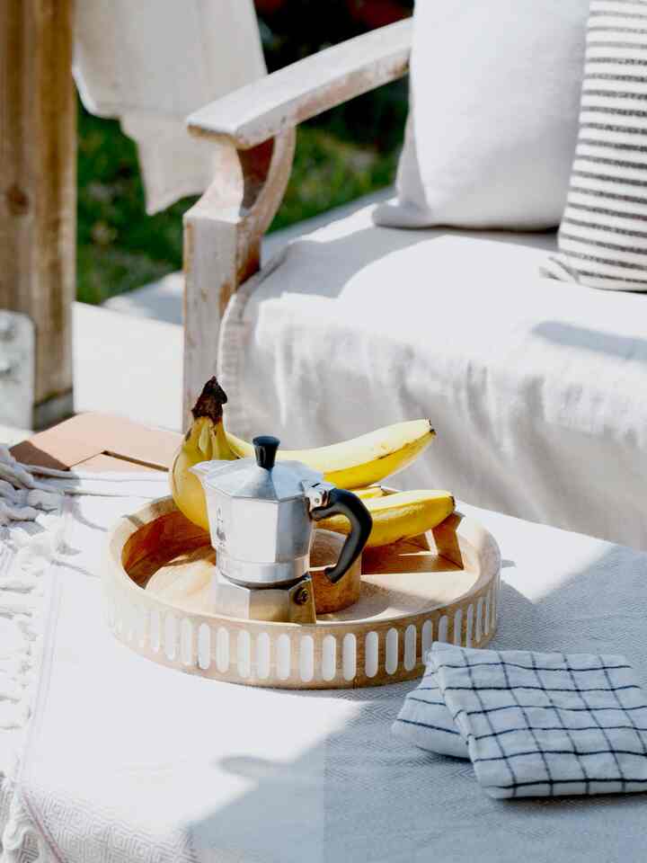 White and wood tone outdoor balcony living space featuring a coffee table with coffee maker and bananas, cozy cushions arranged
