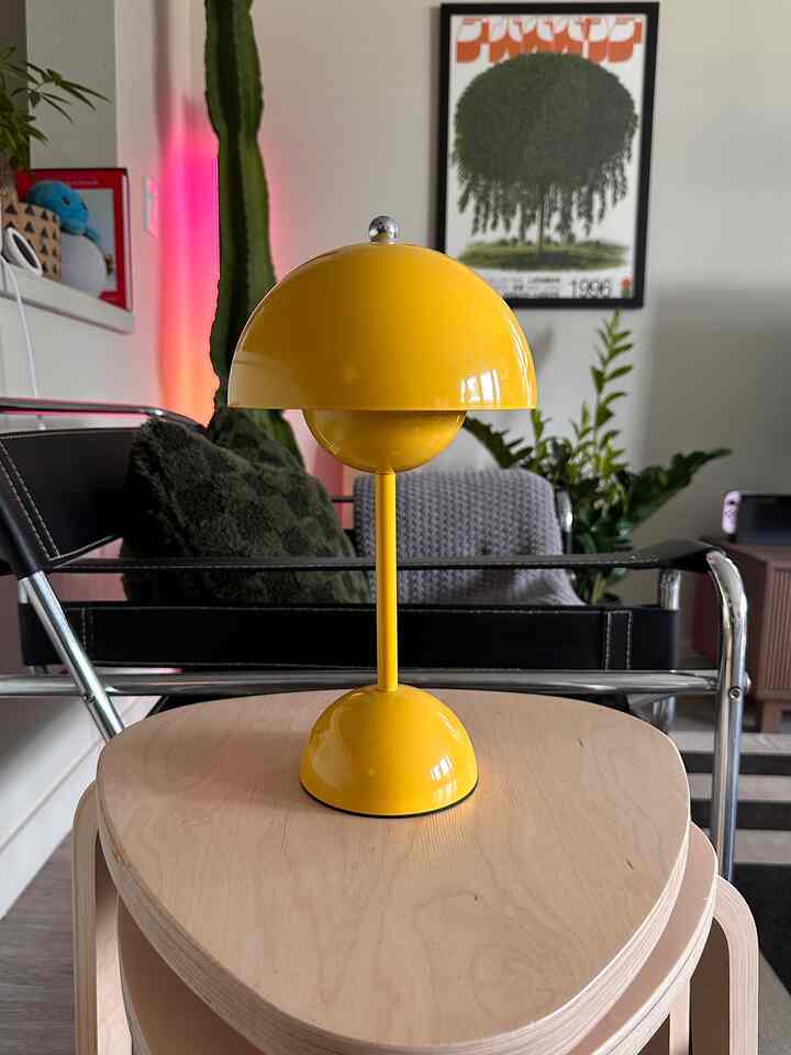 Yellow table lamp on natural wood stools in foreground with black leather chair and multiple plants arranged in a warm living room space