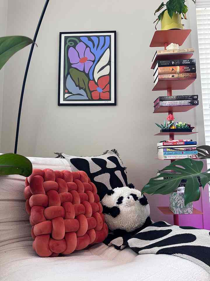 Modern indoor space with beige walls featuring a white sofa, red cushion, bookshelf, and plants arranged cozily