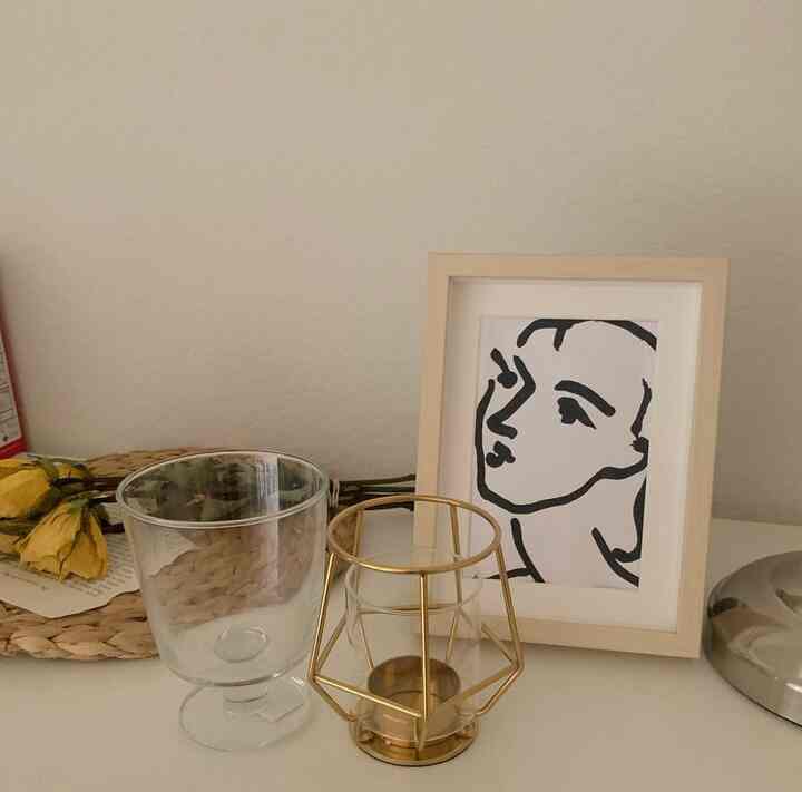 Ivory-toned wall and table feature a gold-frame candle holder, glass, and a framed line art portrait in a neat home gallery setting