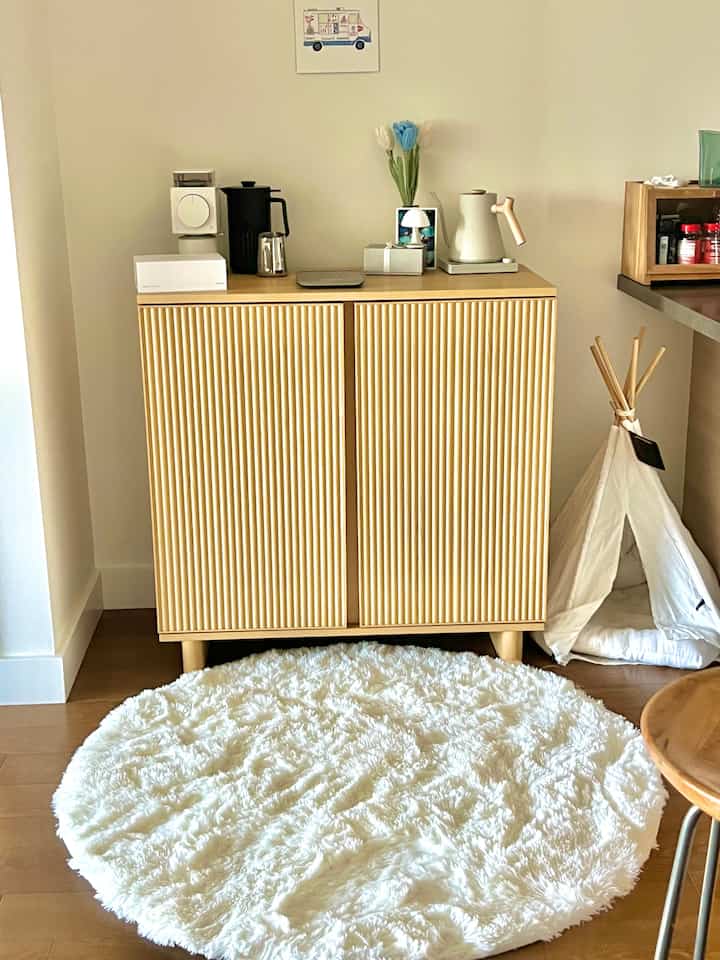 Natural-toned kitchen cabinet area blending white and wood tones, featuring a wooden fluted cabinet, round white faux fur rug, and a pet tent for a cozy vibe