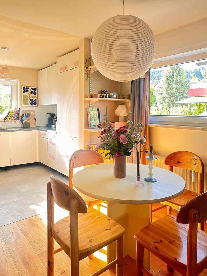 Sunlit kitchen space with warm yellow walls and natural wood tones, featuring a round dining table and wooden chairs, creating a cozy atmosphere