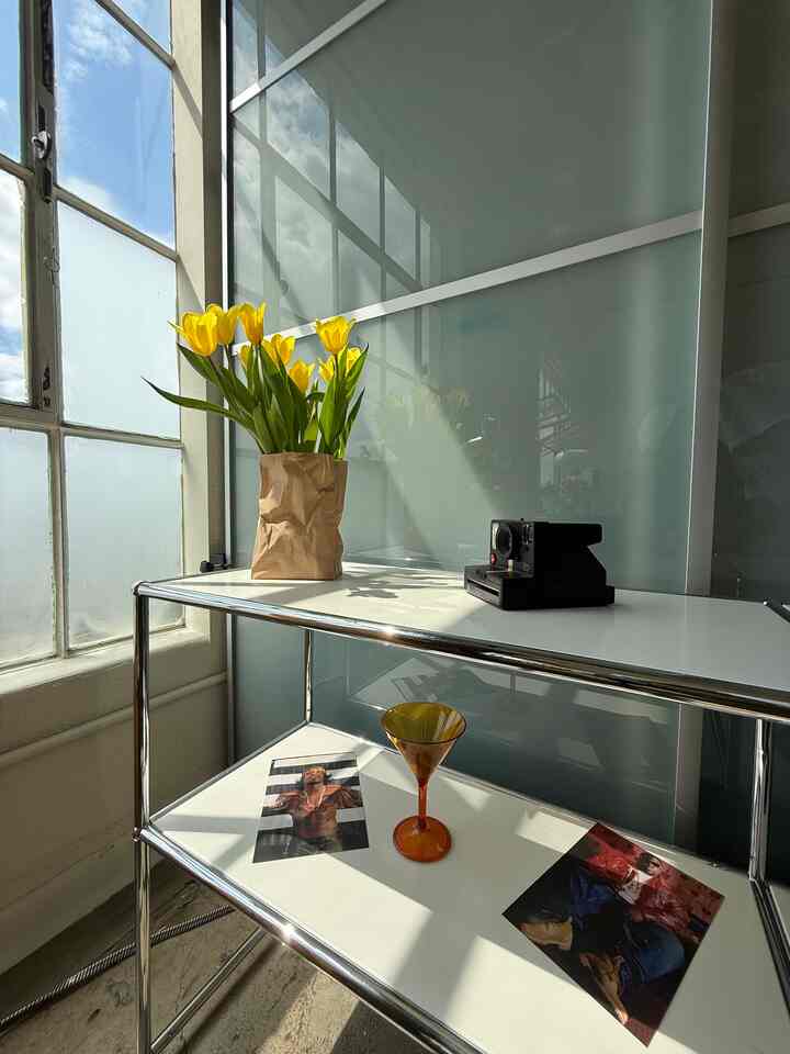 White and yellow tones blend in a stylish space featuring metal shelves by a window with a vase of yellow tulips and vintage camera