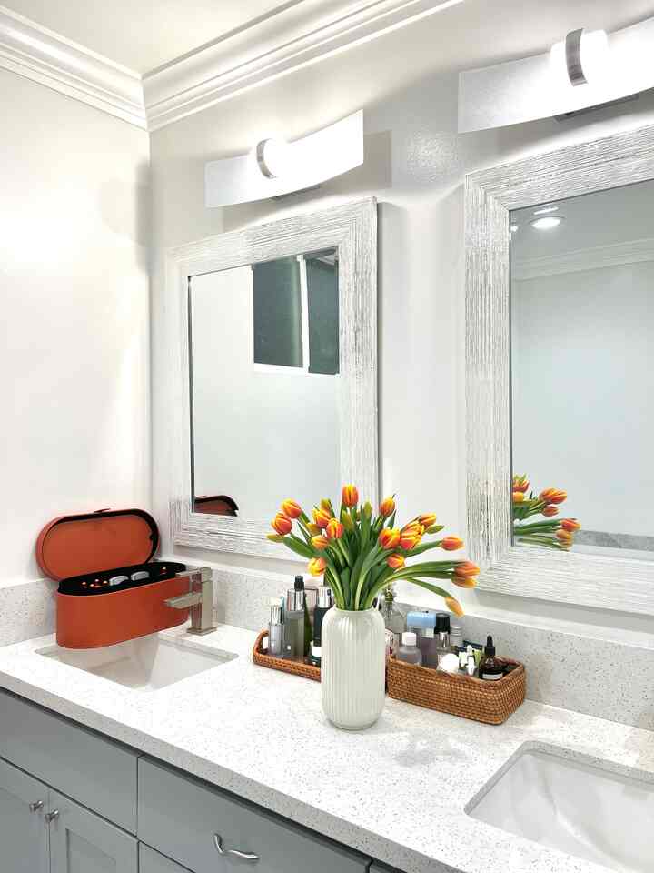 Bright white-toned bathroom vanity featuring two framed mirrors, a white vase with orange tulips, and natural rattan trays creating a fresh, simple ambiance