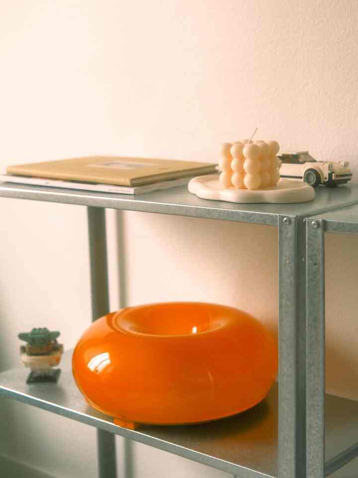 A cozy corner with warm beige walls and metal shelves, featuring a vivid orange donut object, a cute candle, and a miniature LEGO Porsche