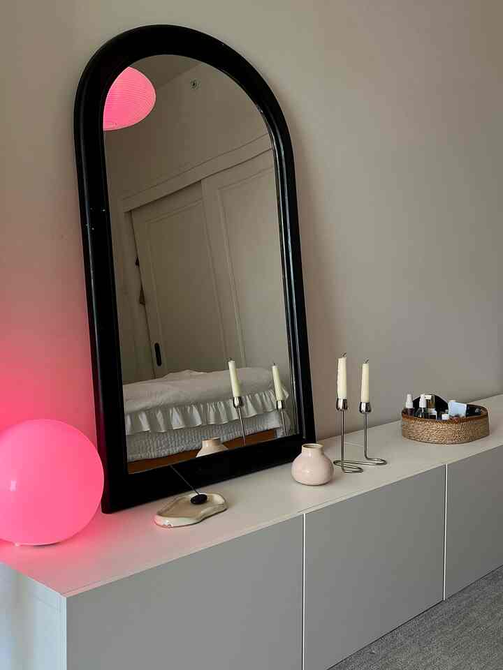 White-toned bedroom featuring a large black arched mirror, white shelf unit, and pink glowing lights creating a cozy mood