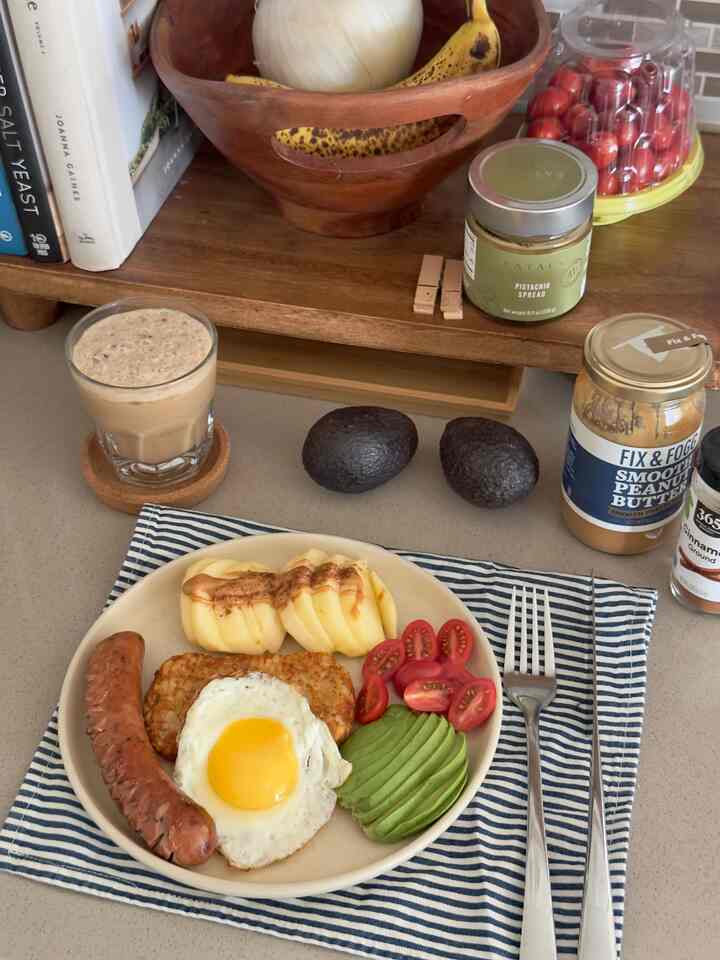 Natural tone kitchen counter featuring breakfast plate with avocado, fried egg, sausage, tomatoes, apple slices, and iced coffee