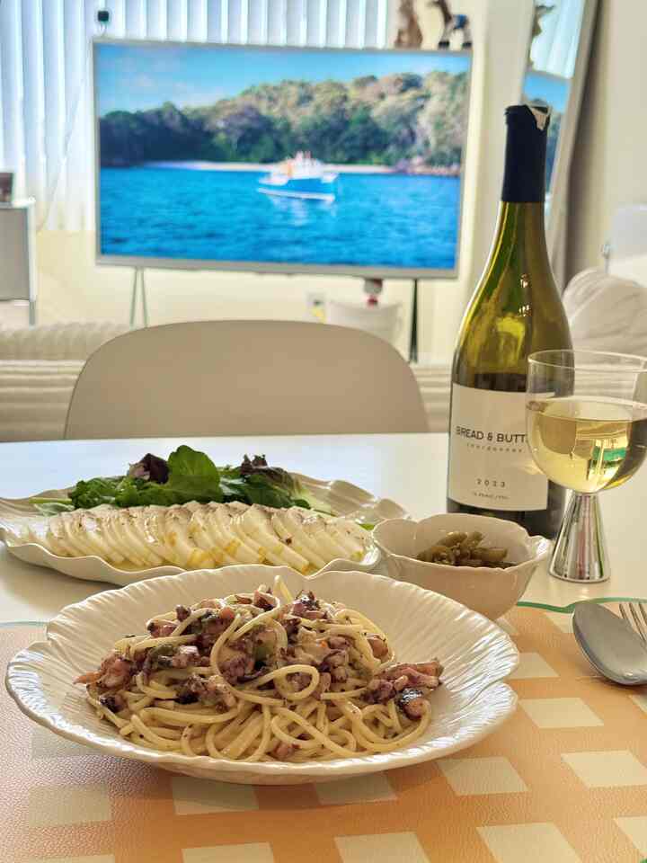White-toned living dining space featuring pasta, salad, wine, and a TV on the dining table, creating a cozy home party atmosphere
