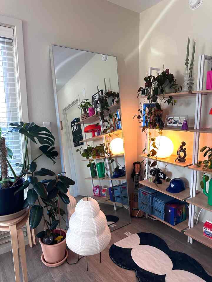 A cozy natural-tone corner space with white and wood tones, featuring corner storage shelves, a large mirror, plants, and a floor lamp.