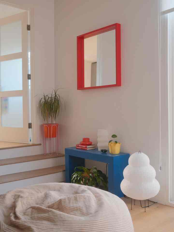Modern natural space with white walls and light wood floor, featuring blue console table, red framed mirror, beige bean bag, and white floor lamp creating a cozy corner