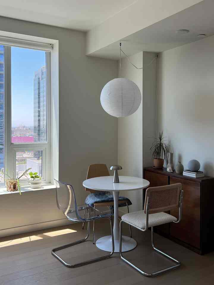 A compact dining room in white and brown tones featuring a round dining table with transparent chairs and minimalist lighting in a simple setting