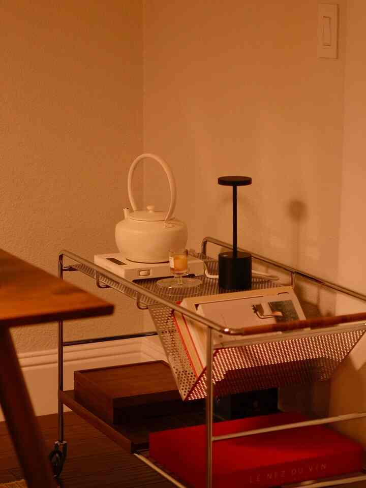 Warm-toned dining room featuring a bar cart with white kettle, black table lamp, magazines, and decorative items arranged harmoniously