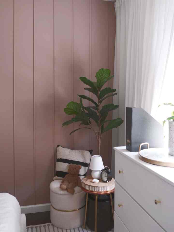 A cozy bedroom corner in warm beige and white tones featuring an ottoman with cushions and a green plant accented by soft drapes
