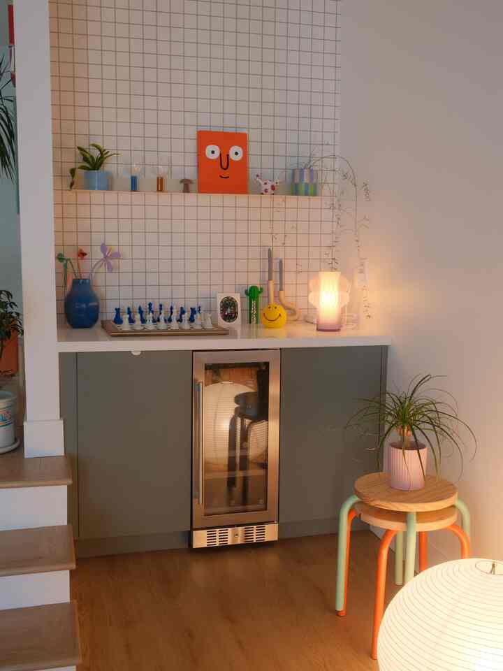 Bright white tiled wall corner storage with wood floor, featuring vases, plants, and a table lamp creating a cozy atmosphere