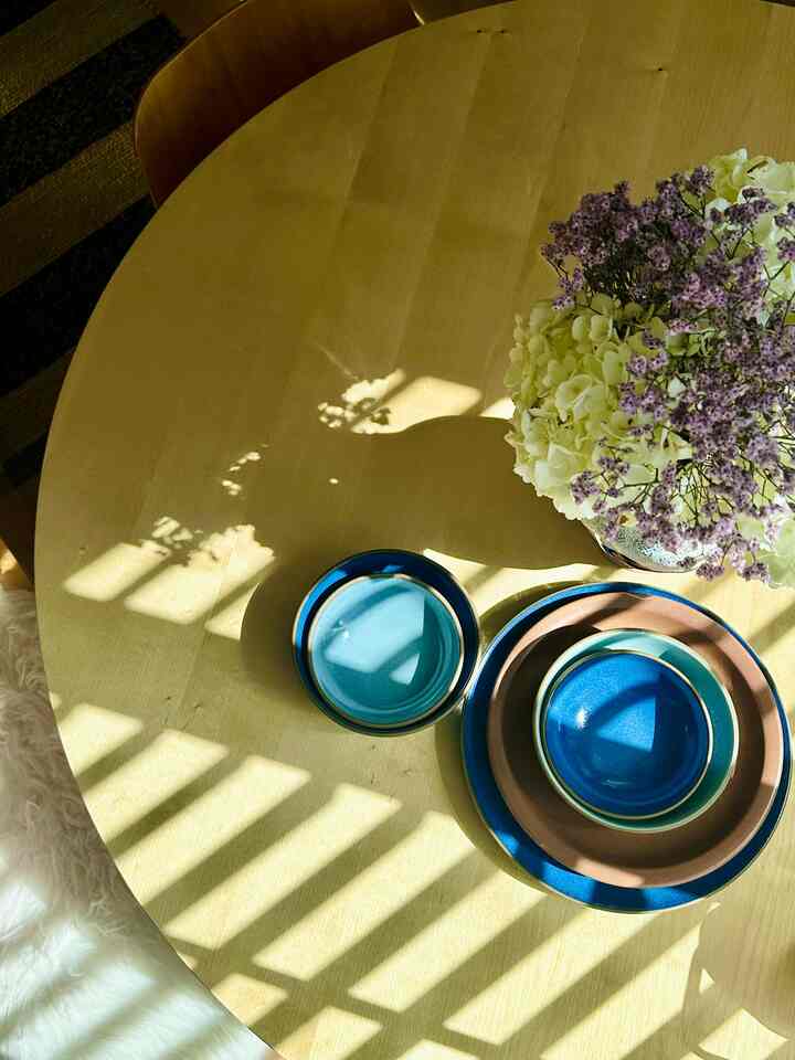 Natural-toned round dining table featuring blue and brown plates and bowls with a vase of purple and white flowers arranged on top