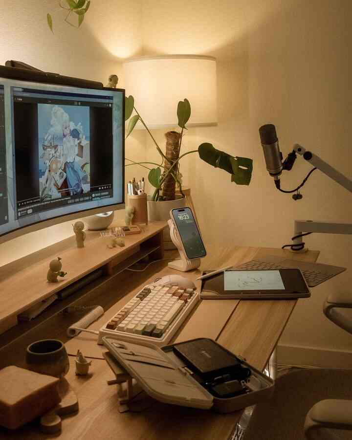 A natural wood-tone home office featuring a curved monitor, microphone, and plant with warm ambient lighting creating a cozy workspace