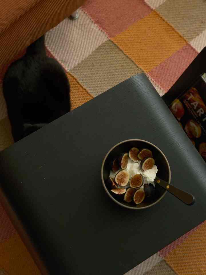 Black and orange toned rug with a black table and black cat beside it; table holds bowl of figs and cream, creating a warm home cafe atmosphere