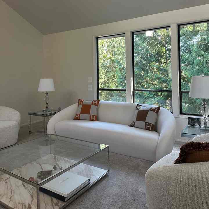 Bright and clean white living room featuring a white sofa with orange cushions, glass coffee table, and clear table lamps with natural greenery through large windows