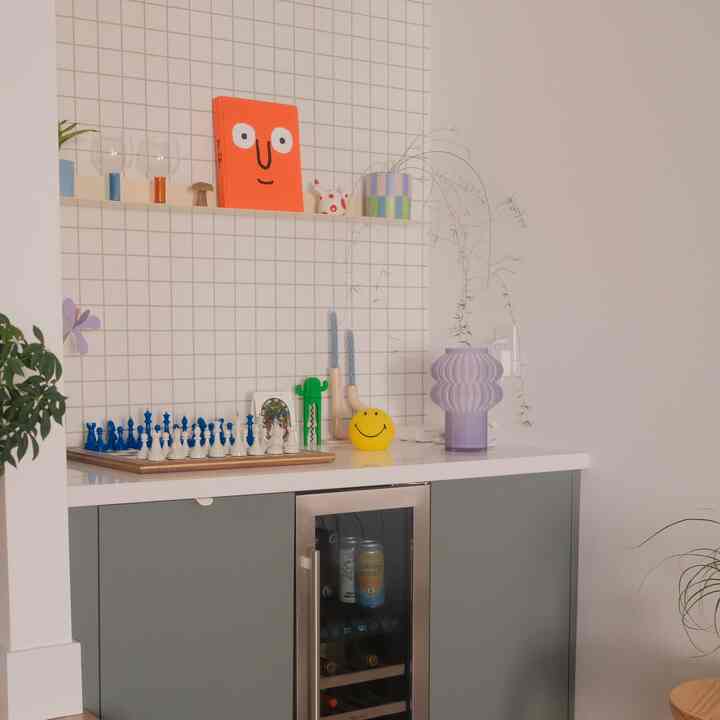 Home bar corner with white tiled wall and green lower cabinets, featuring a purple table lamp and a chess set, creating a cozy atmosphere