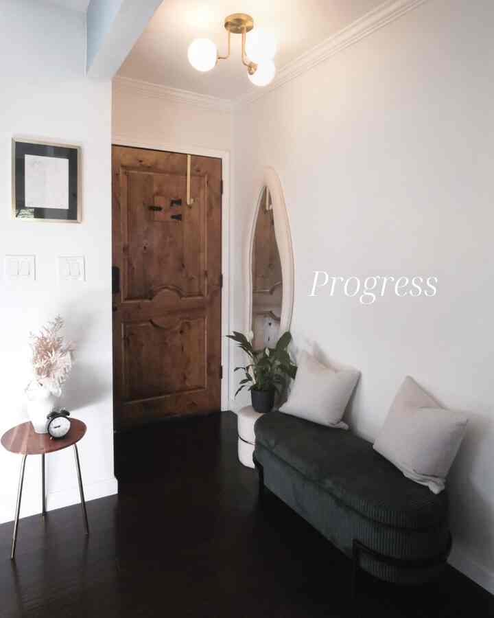 Bright white-walled entrance featuring a wood-tone door, storage bench with cushions, oval mirror, and a potted plant for a cozy modern feel