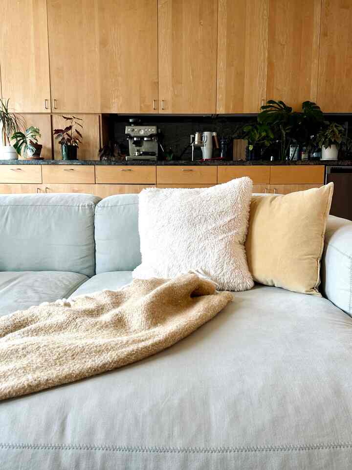 Natural color and beige toned living room featuring a cozy light blue sofa with cushions and wood-toned kitchen cabinetry