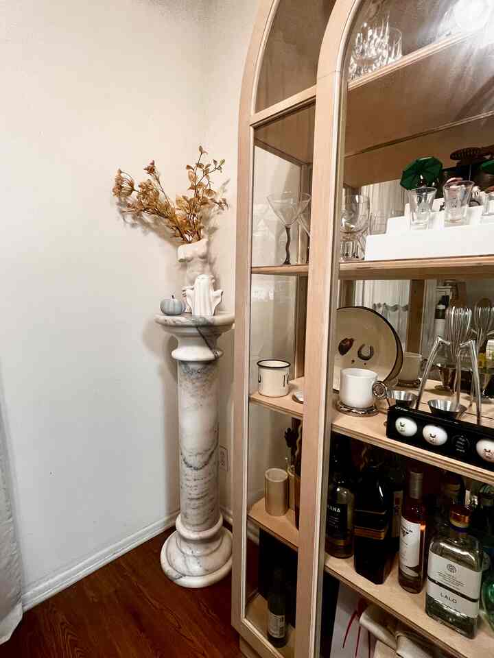 Natural tone living room corner featuring a marble pedestal with a vase and dried branches, wood furniture, and a cozy home bar setup