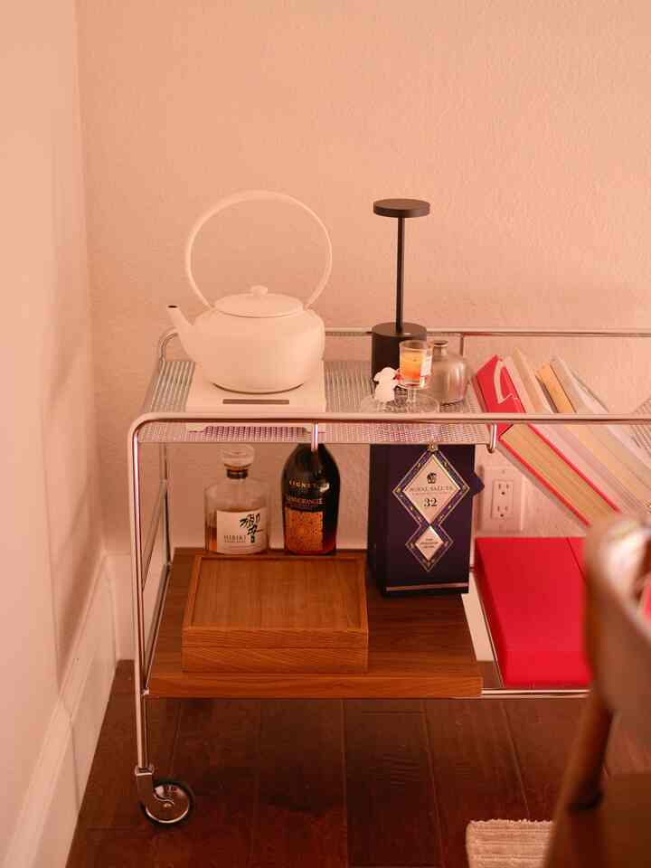 A brown wood floor corner of a dining room with a metal and wood bar cart featuring a white teapot and candle on top against a white wall background