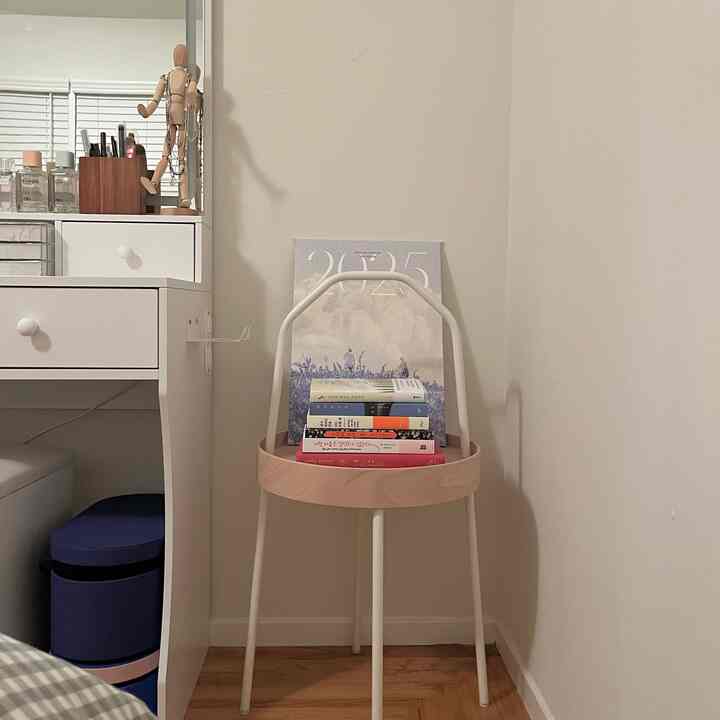 White and brown toned bedroom corner featuring a simple white desk and a small bookshelf with stacked books, creating a cozy reading nook