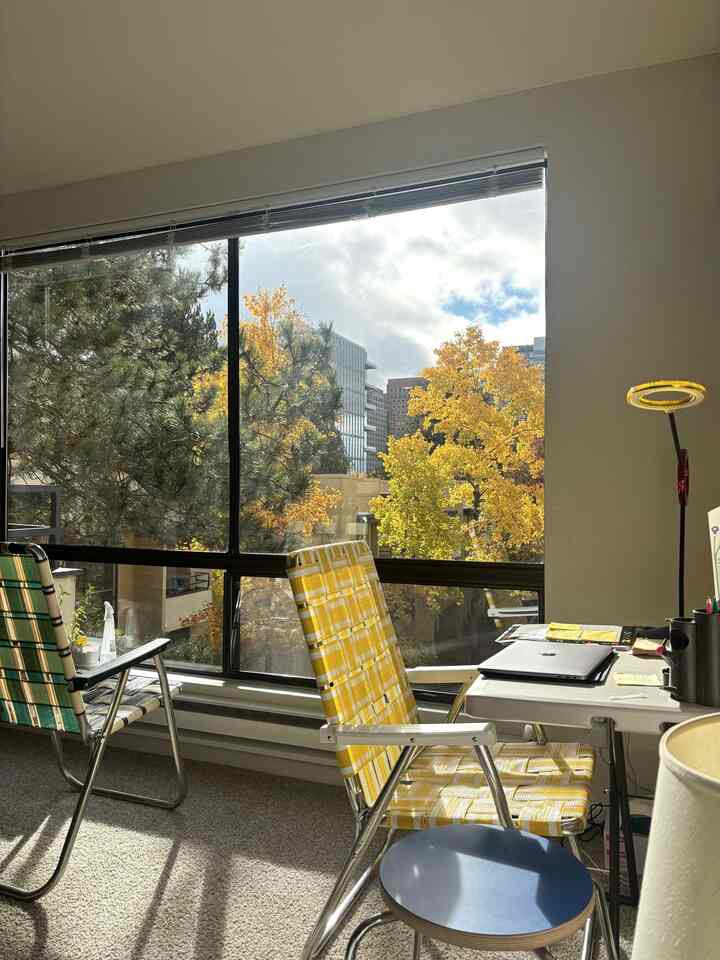 A naturally lit home office and casual dining space featuring yellow and brown fall foliage visible through large windows with stools and woven chairs