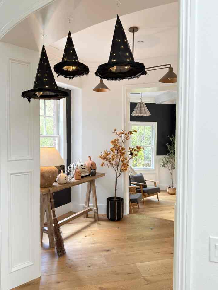 White-walled entrance featuring wood-tone flooring, illuminated hanging witch hats as Halloween lighting, rustic reclaimed wood console table with Halloween pumpkins