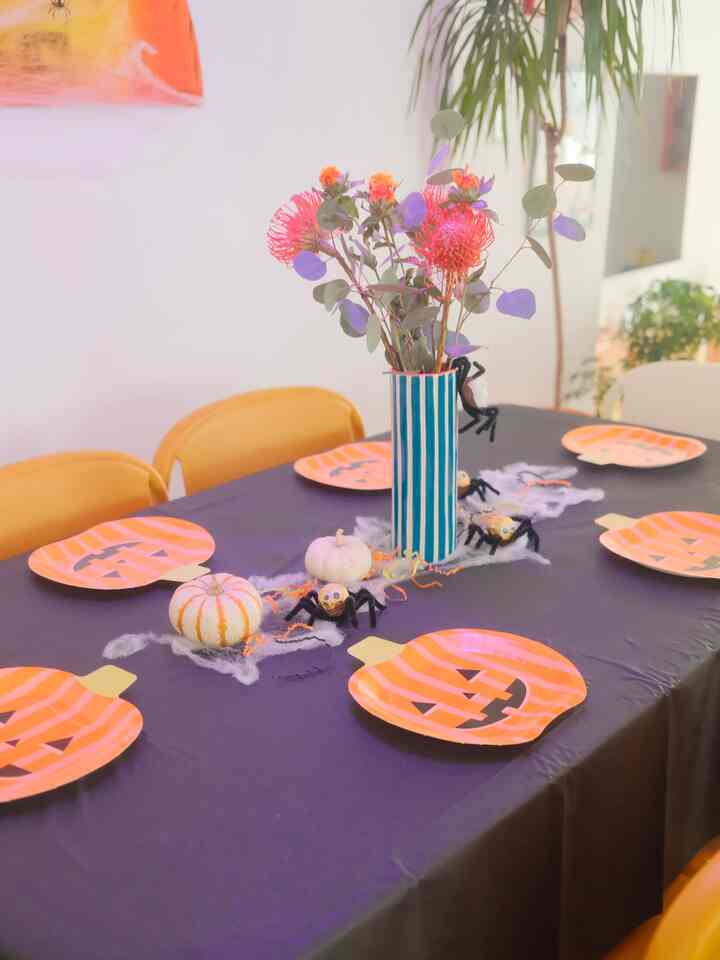 Black covered dining table decorated with orange pumpkin plates and floral vase centerpiece, surrounded by mustard yellow chairs for a Halloween party vibe