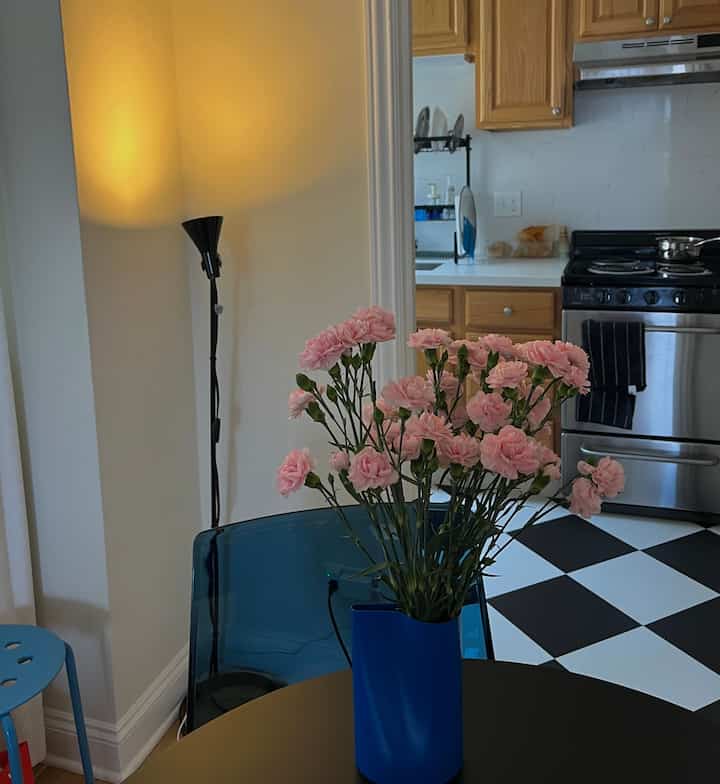 Cozy kitchen and dining area with warm lighting, pink flowers in blue vase, natural wood cabinets, and checkerboard floor