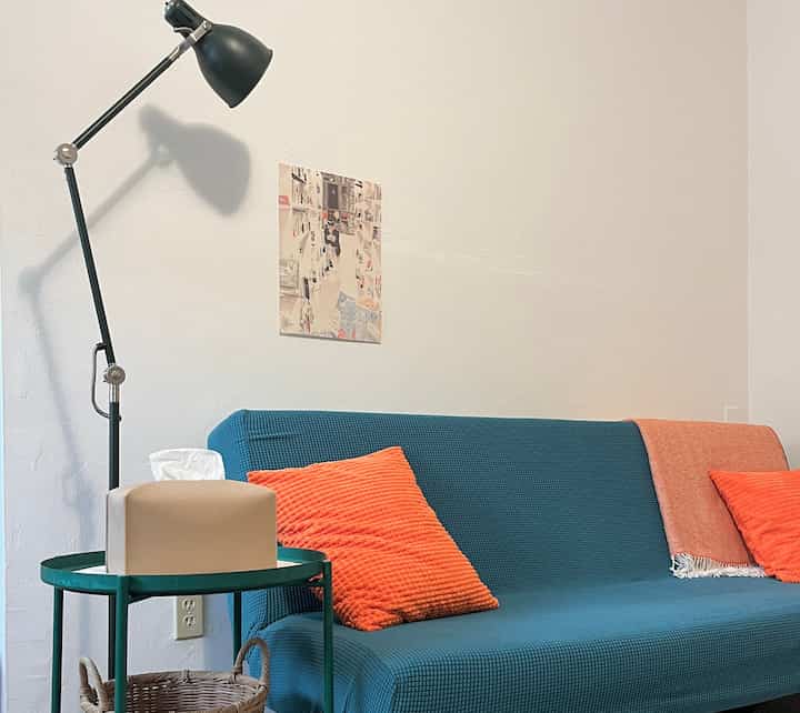 Living room with white walls and a blue sofa, accented by orange cushions and a floor lamp creating a simple, cozy atmosphere