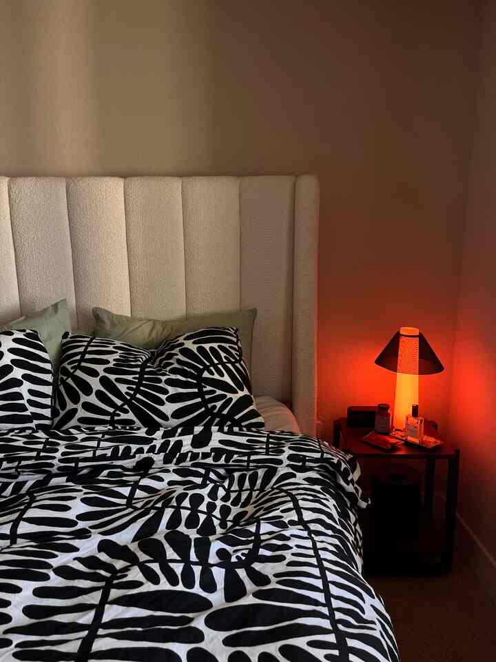 Beige-toned bedroom featuring a white upholstered headboard bed with black leaf patterned bedding, red-lit table lamp on nightstand