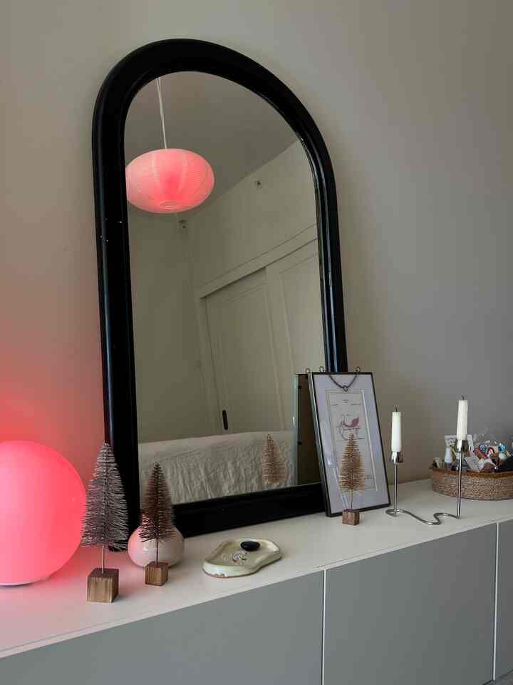 White and black toned bedroom featuring an arched black mirror and white cabinet topped with miniature Christmas trees and soft pink lamp, minimal decor