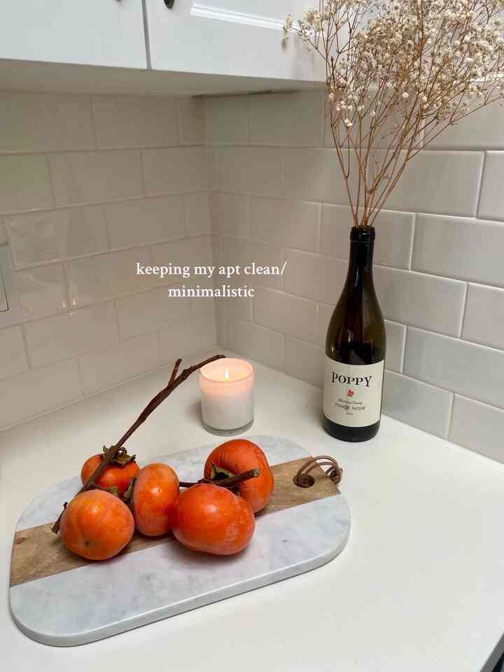 Clean, minimalistic kitchen corner with white tiled walls, marble tray holding persimmons, candle and dried flowers