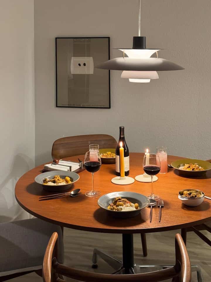 Minimal dining room with brown wooden round table and pendant light, cozy home party atmosphere enhanced by candles and wine glasses