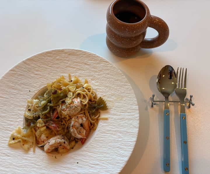A simple one-person dining setup on a white table featuring a brown mug and sky blue cutlery with a pasta plate