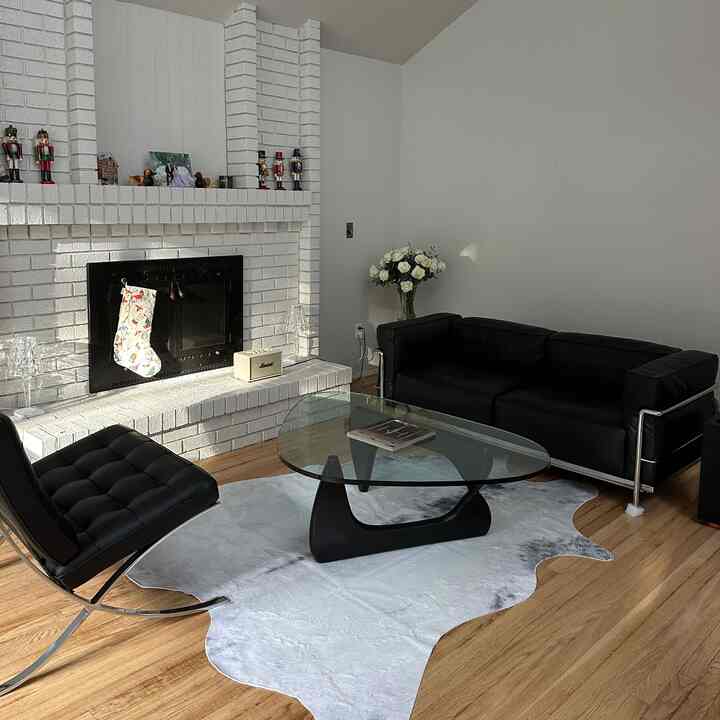 Modern living room featuring white walls and fireplace, black leather sofa and armchair, glass coffee table, and wood-tone flooring