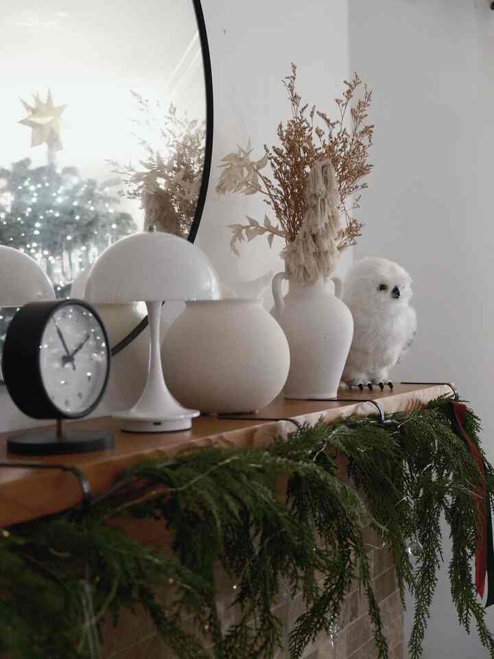 Cozy holiday-decorated living room fireplace mantel in white and brown tones featuring mirror, table lamp, vases, and owl objet