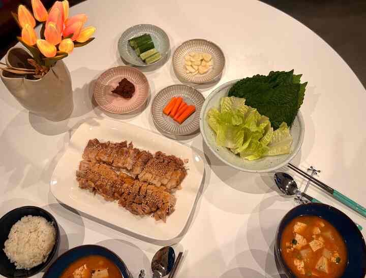 White-toned dining area featuring a table set with bossam and doenjang-jjigae, alongside a vase with flowers creating a cozy atmosphere