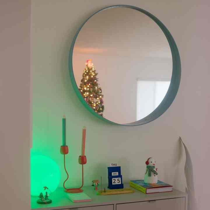 A white-walled space with a STOCKHOLM round mirror reflecting a decorated Christmas tree and festive ornaments creating a cozy vibe