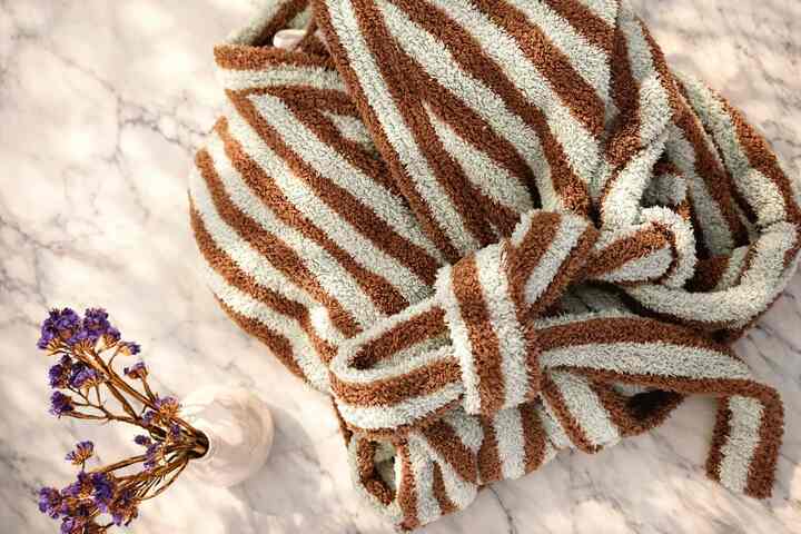 Brown and white striped shower robe on white marble surface with small vase of dried purple flowers, creating a clean and warm bathroom accessory scene