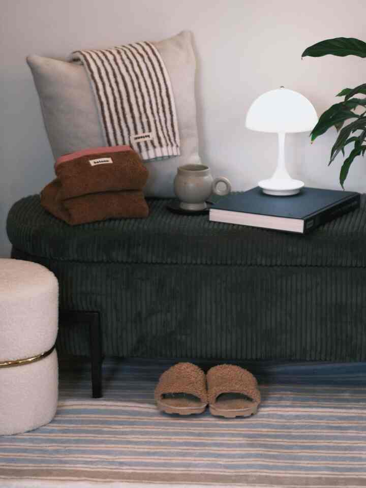 Natural-toned entrance space featuring a green ottoman with a table lamp, mug, and towels creating a cozy atmosphere