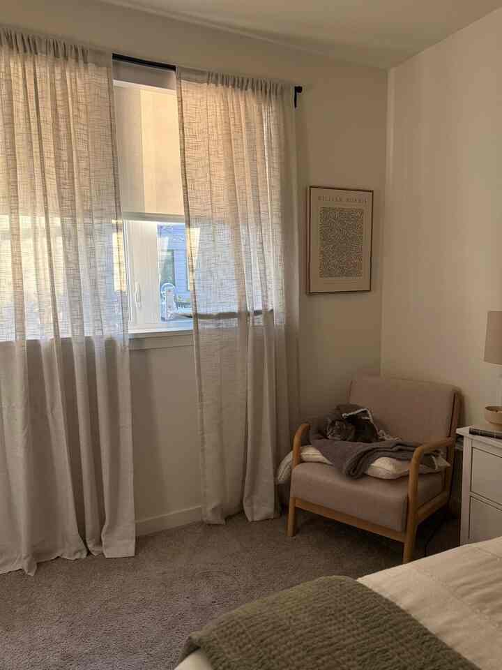 Cozy beige-toned bedroom corner featuring an armchair with a resting cat on blankets and curtains softly filtering sunlight from the window