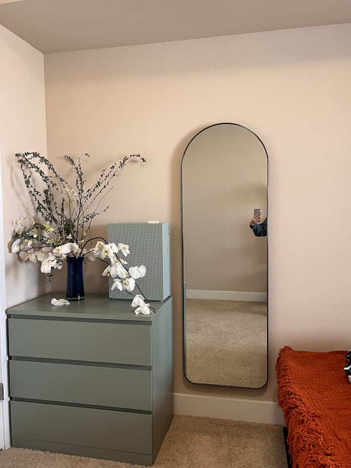 Natural tone cozy interior featuring a green three-drawer cabinet and arched mirror against beige walls and white carpet flooring