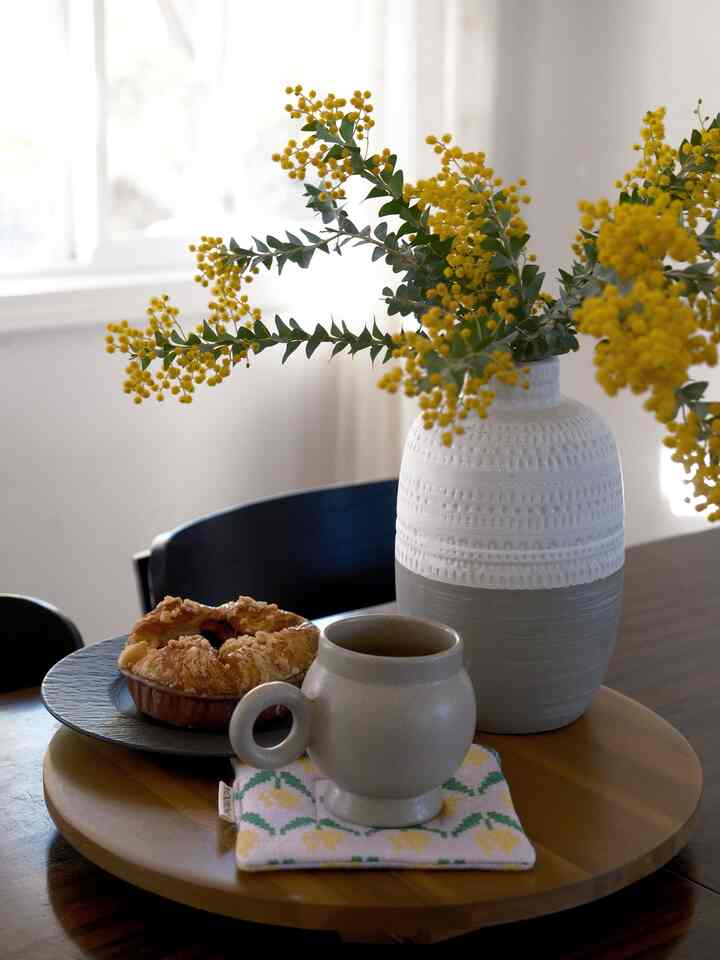 Modern dining room with warm wood-tone table featuring a yellow flower vase and croissants