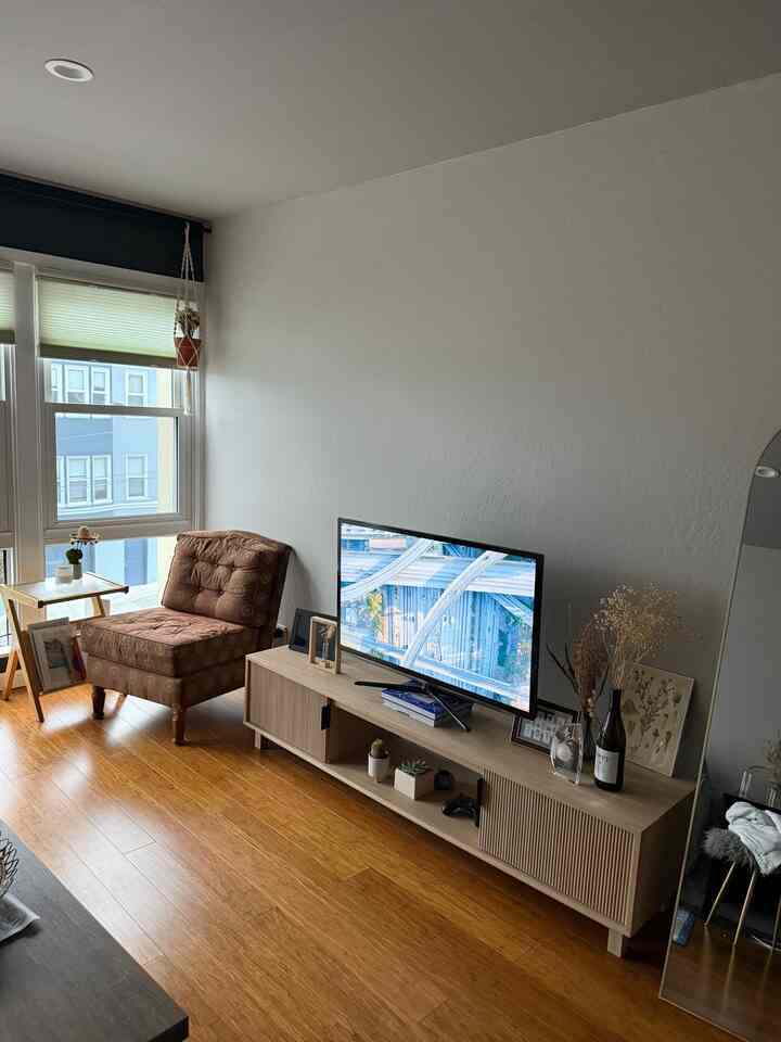 Living room with wood tone flooring and white walls, medium sized TV stand and brown patterned sofa chair, featuring a natural ambiance